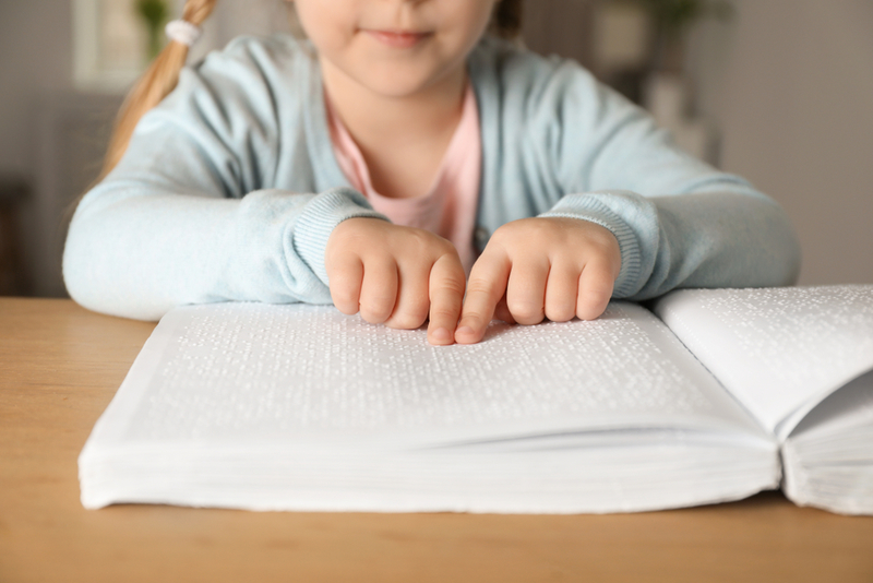 Foto de uma criança lendo um livro em braile. A criança está de frente, É possivel ver o rosto da criança do nariz para baixo. Ela toca com os dois dedos indicadores a página de um livro branco que está sobre a mesa.